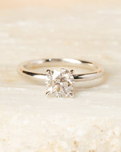 Load image into Gallery viewer, A classic solitaire engagement ring with a round brilliant-cut diamond set in a polished silver-colored band, resting on a textured light beige stone against a soft beige background.