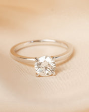 Load image into Gallery viewer, A classic solitaire engagement ring with a round brilliant-cut clear gemstone in a four-prong setting on a polished silver band, resting on draped beige fabric.
