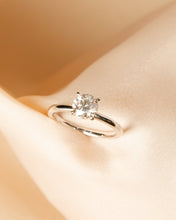 Load image into Gallery viewer, A silver solitaire engagement ring with a round-cut clear gemstone, displayed on soft draped beige satin fabric.