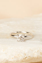 Load image into Gallery viewer, A classic solitaire engagement ring with a round brilliant-cut diamond set in a polished silver-colored band, resting on a textured light beige stone against a soft beige background.
