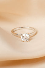 Load image into Gallery viewer, A classic solitaire engagement ring with a round brilliant-cut clear gemstone in a four-prong setting on a polished silver band, resting on draped beige fabric.
