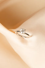 Load image into Gallery viewer, A silver solitaire engagement ring with a round-cut clear gemstone, displayed on soft draped beige satin fabric.
